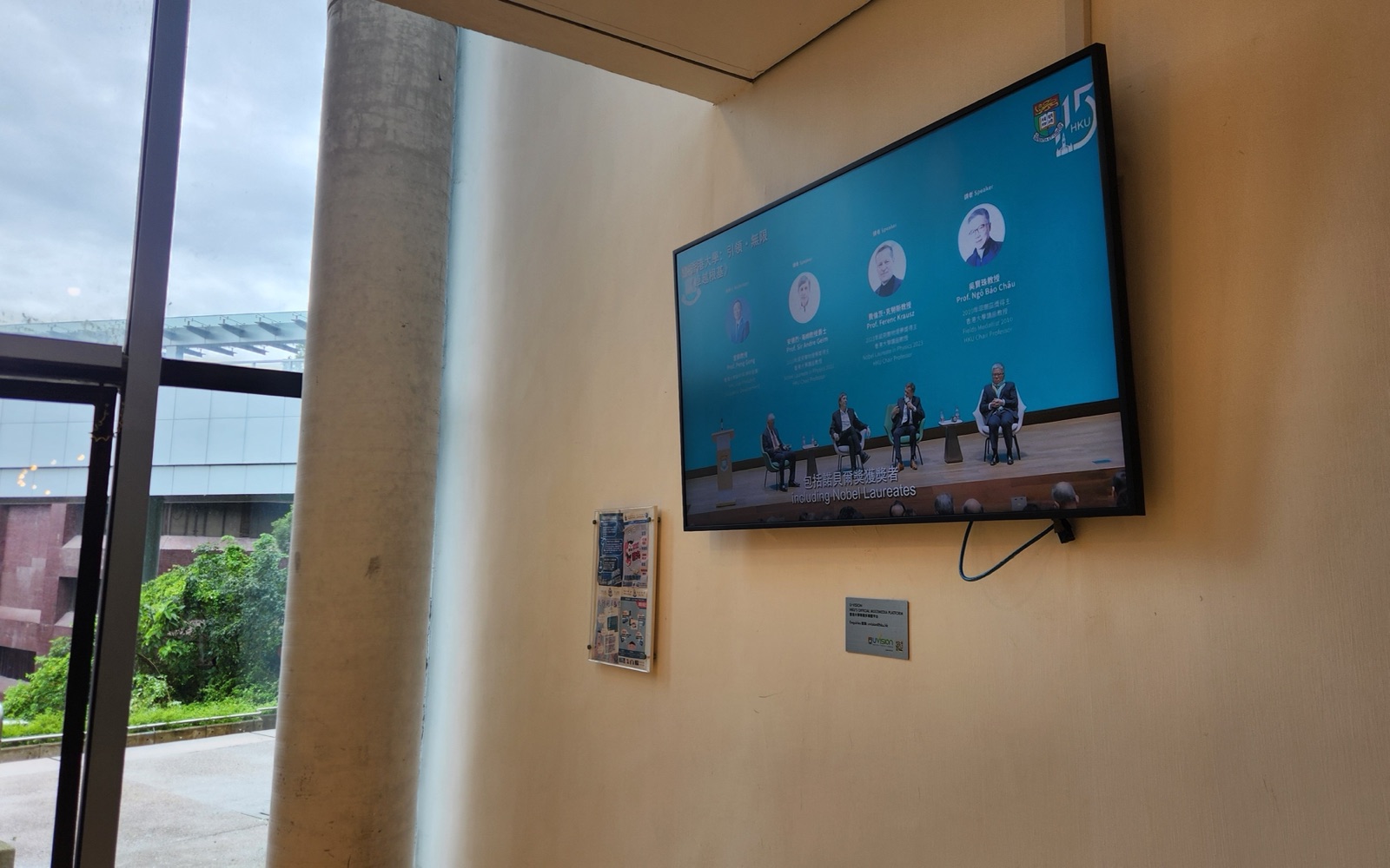 A U-Vision screen mounted in the glazed corner of the Graduate House lobby, overlooking the courtyard